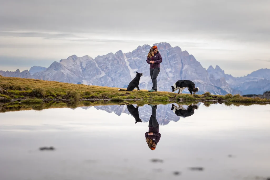 Wunderschönes Bild von Mensch mit spielenden Hunden vor einem kleinen See. Im Hintergrund die mächtigen Berge der Dolomiten.